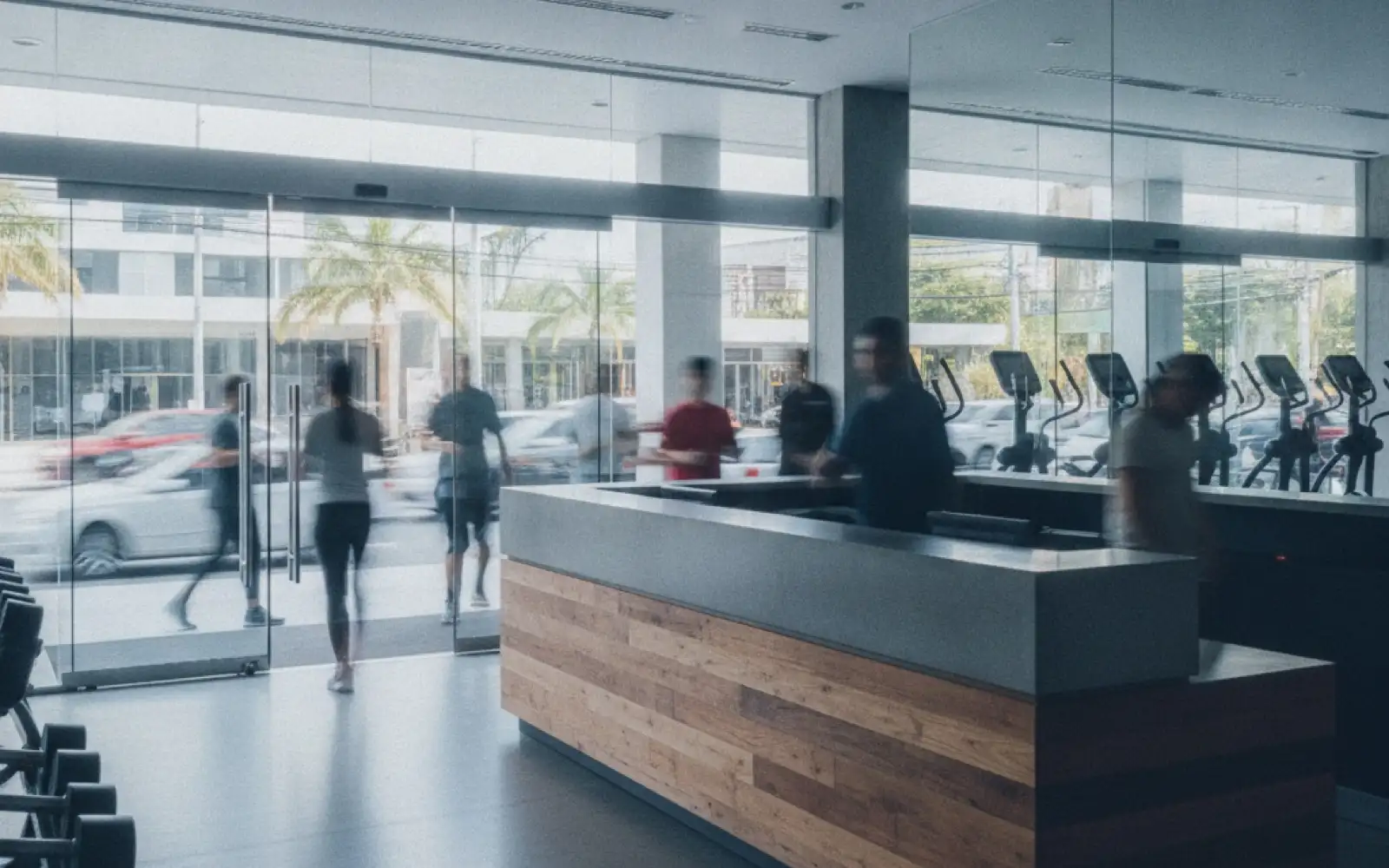 Recepción de un gimnasio con mostrador de madera y gente entrando por la puerta de cristal.